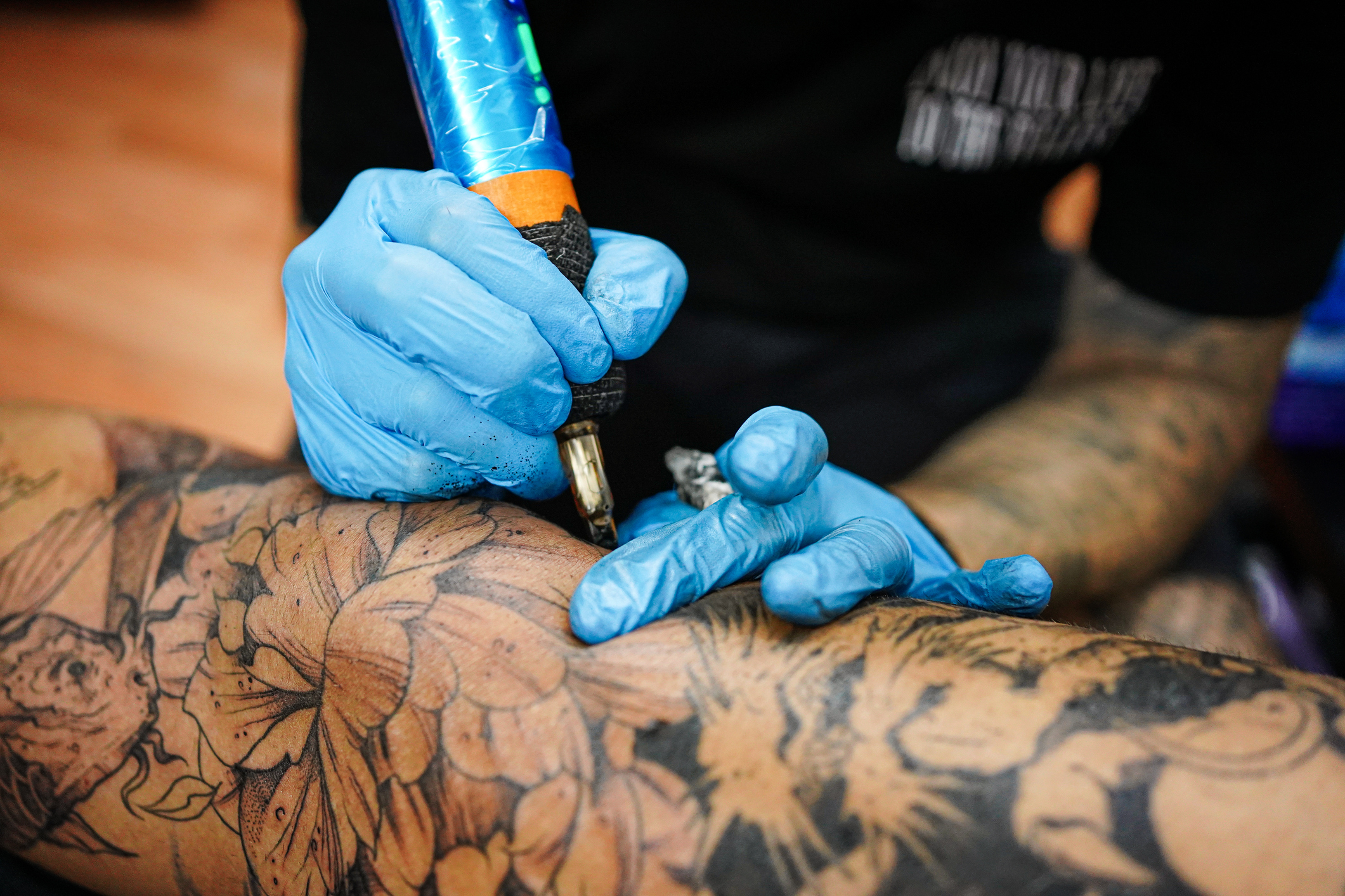 A close-up shot shows a tattoo artist's gloved hands working on a large, intricate tattoo on a person's leg. The artist is wearing blue disposable gloves and holding a tattoo machine with a blue and orange grip. The machine's needle is touching the skin, adding to a detailed floral and animal design that covers a significant portion of the leg. The existing tattoo work appears to be in various stages of completion, with some areas fully shaded in black and others still in outline form. The background is blurred, suggesting a focus on the tattooing process.